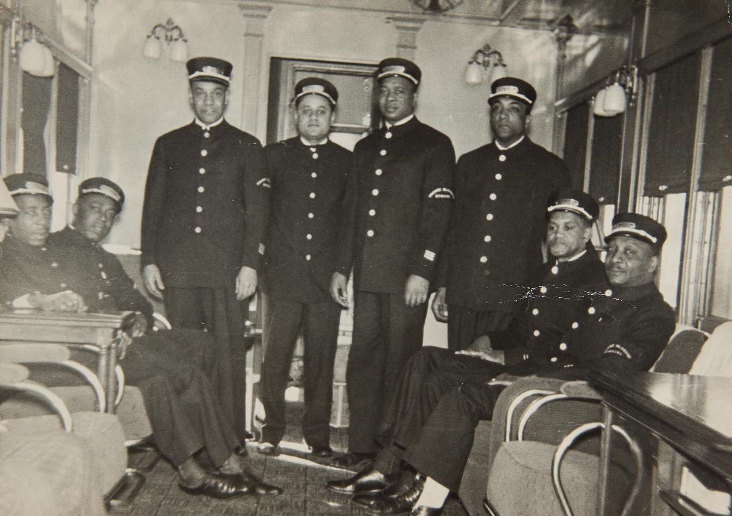 A black and white photograph of eight members of the Brotherhood of Sleeping Car Porters, all in uniform, sitting or standing in a train cabin. Photo Source: The National A. Philip Randolph Pullman Po