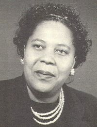 A black and white photograph of Smith, who is wearing a suit and pearl or shell necklaces and earrings looking up and away from the camera with a soft smile. Photo Source: FTA