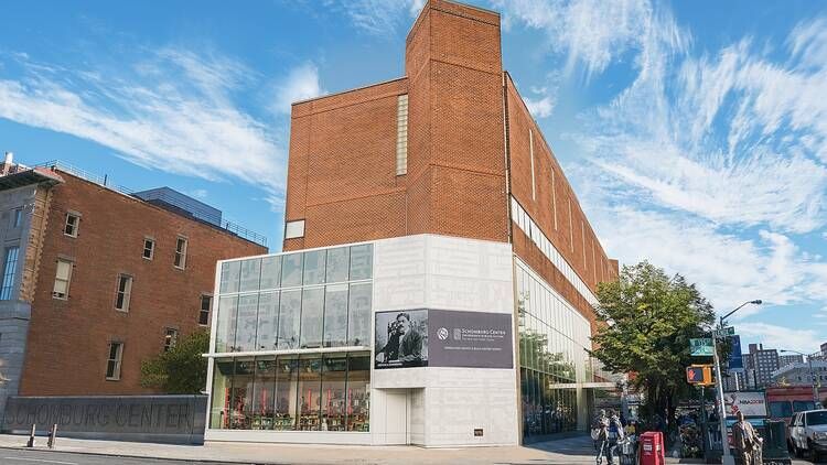 A photograph of the Schomburg Center from across the street against a blue sky with clouds. On the face of the building is a banner with a photograph of Arturo Alfonso Schomburg. Photo Source: Schombu