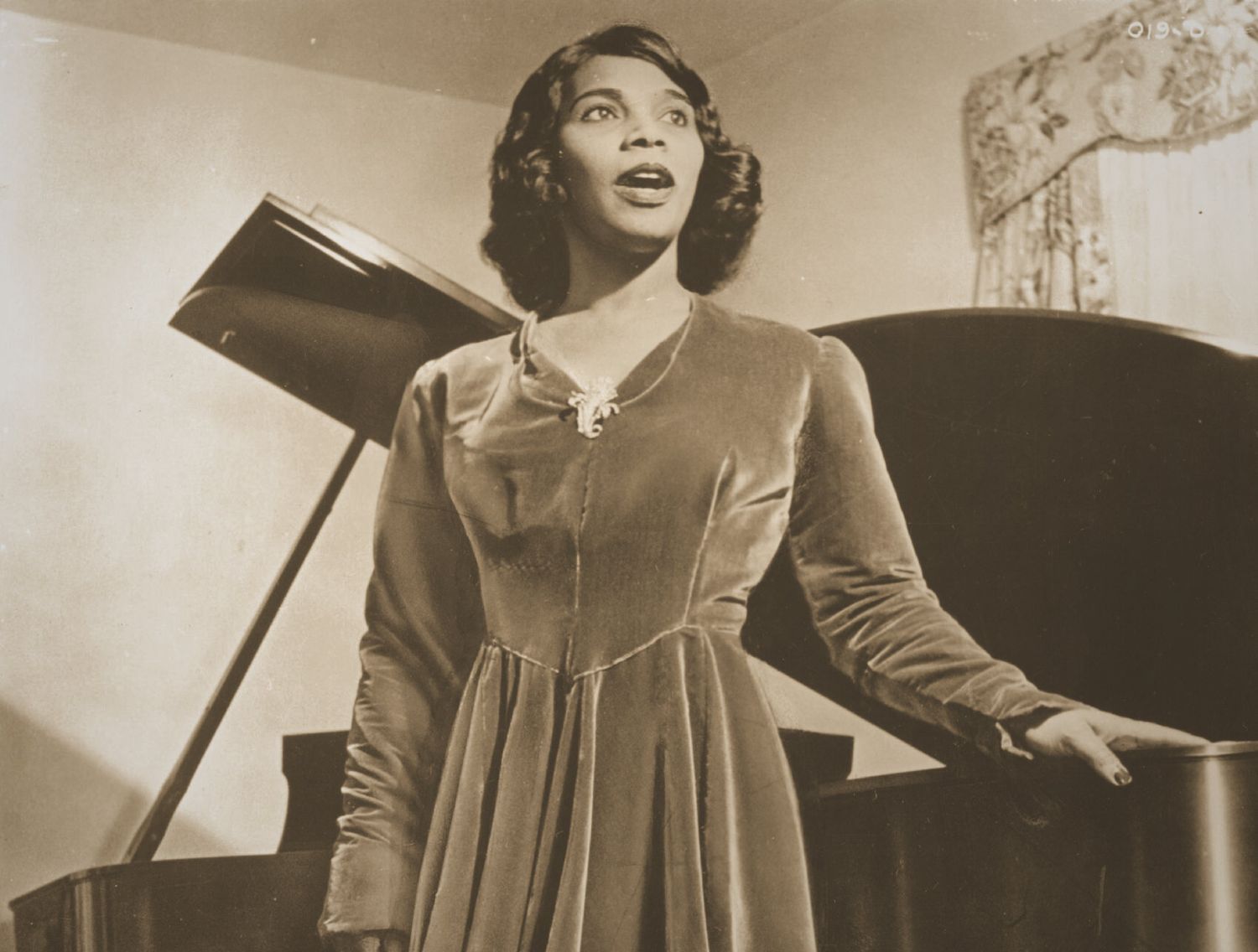 A sepia toned photograph of Anderson, standing against a grand piano, wearing a velvet dress and singing, circa 1935. Photo Source: Michael Ochs Archives/Getty Images