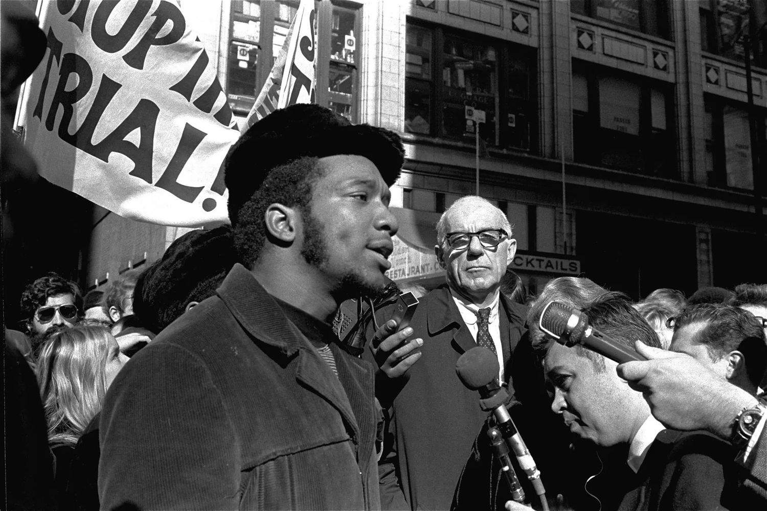 A black and white photograph of Fred Hampton, center, speaking to reporters at a rally outside the federal courthouse in Chicago on Oct. 29, 1969. Doctor, athlete and activist Benjamin Spock is on his