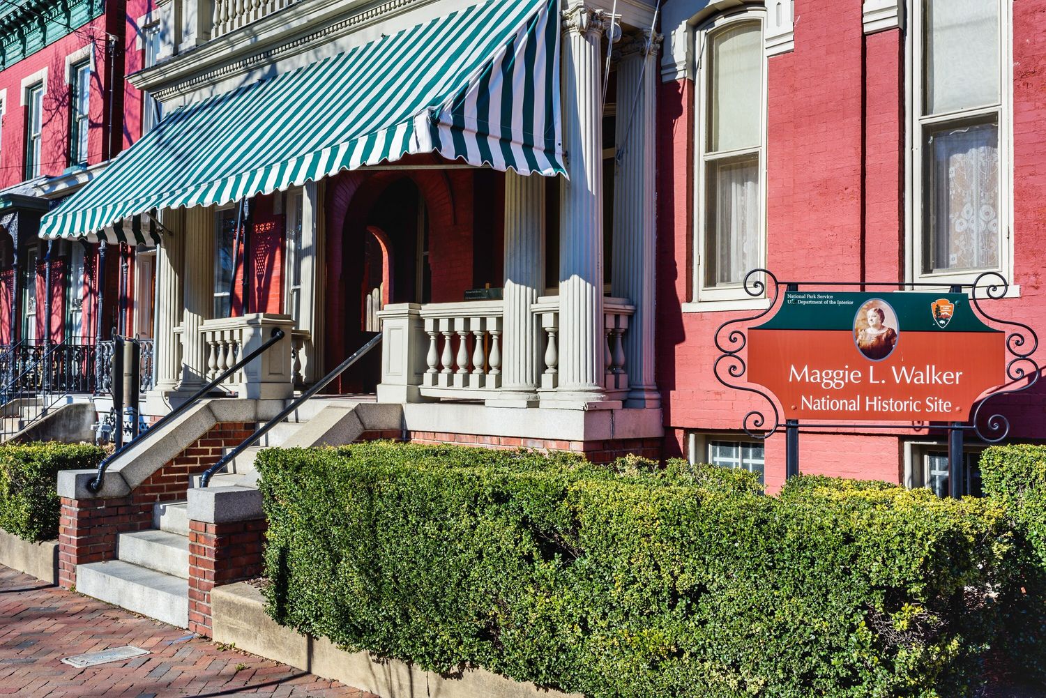 A photograph of the exterior of Walker’s home as part of the Maggie L. Walker National Historic Site, bathed in full sun with well manicured hedges and a clean sidewalk. Photo Source: National Park Se
