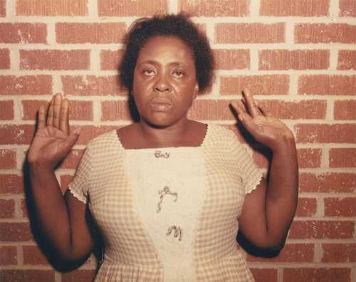 A color photograph of Hamer against a brick wall, hands up, with a defiant look on her face, after being beaten in jail in 1964. Photo Credit: FBI.