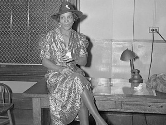 Black and white photograph of St. Clair seated atop a desk, wearing a fedora and a sparkly dress. Photo Source: Blackpast.
