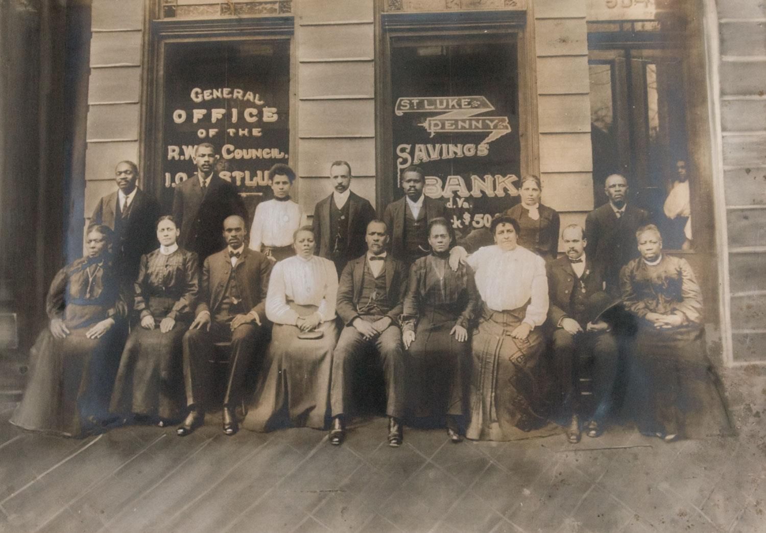 A black and white photograph Walker (front, left of center) with over a dozen of her employees, seated for a formal portrait in two rows outside the St. Luke Penny Savings Bank. Photo Source: virginia