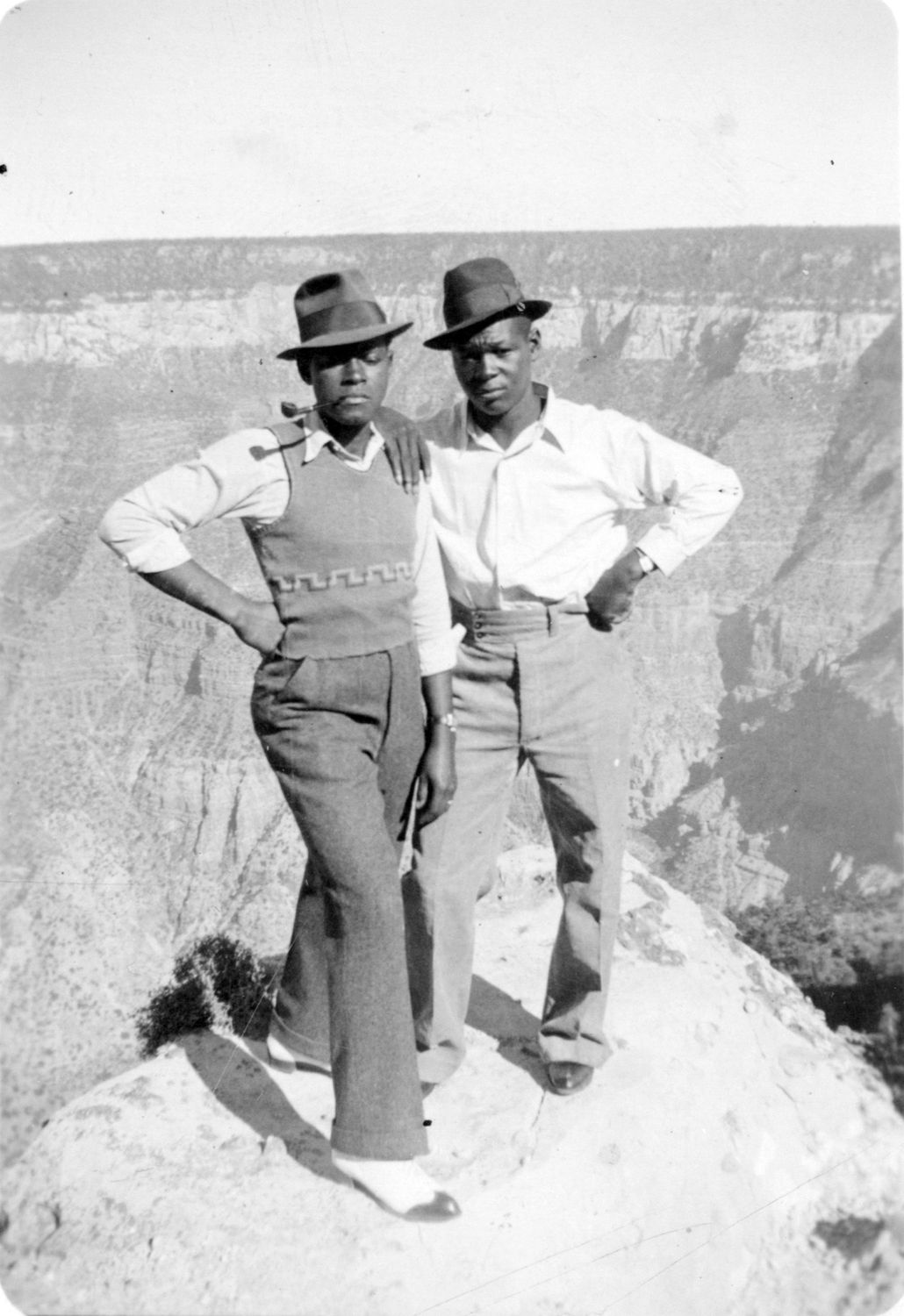 A black and white photograph of two Black men, dressed sharply, standing on the edge of Grand Canyon in nice pants and fedoras, taken by John B. Scott. Photo Source: Grand Canyon NP Museum Collection
