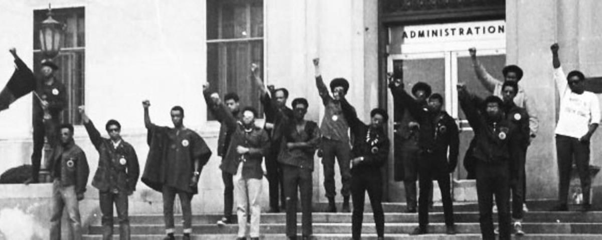 A black and white photograph of a group of seventeen Black United Students standing on the steps of the university’s administration with their fists raised in Black solidarity. Photo Source: Kent Univ