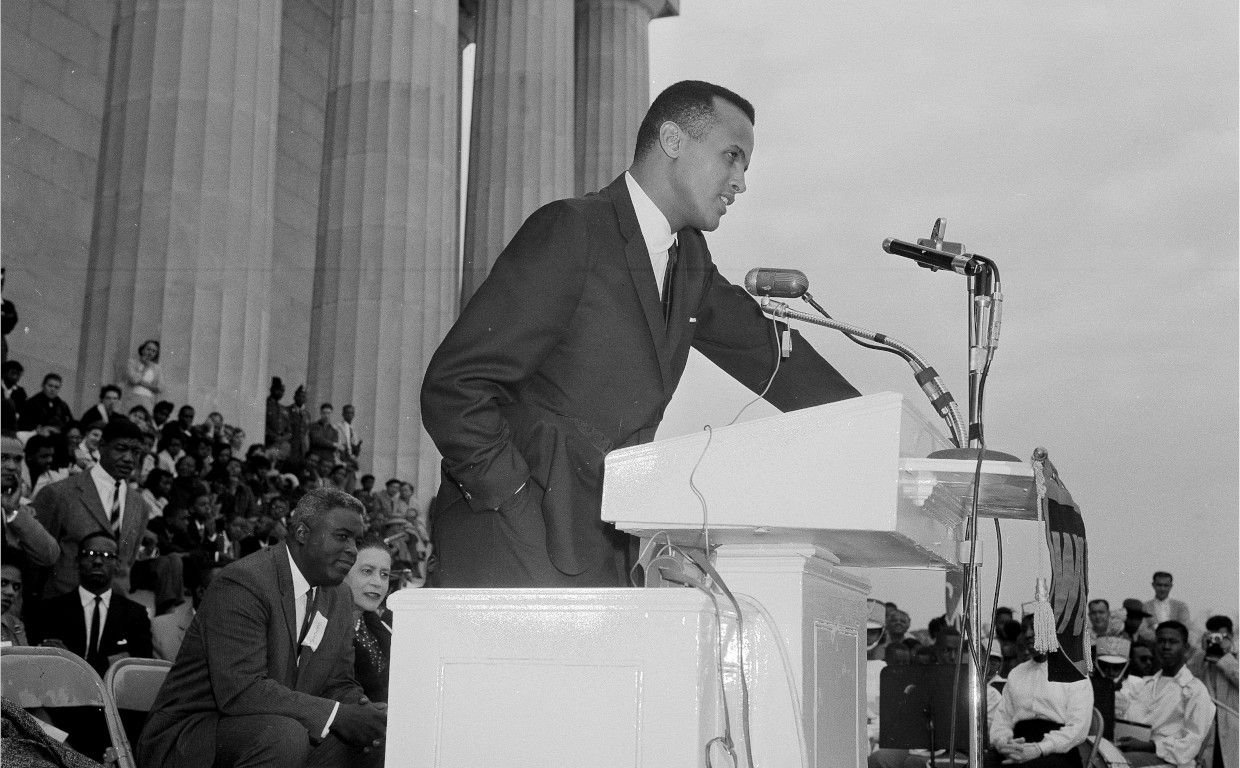 Harry Belafonte speaking at a lectern at the Youth March for Integrated Schools. Jackie Robinson can be seen sitting behind him at left. Photo Source: Associated Press