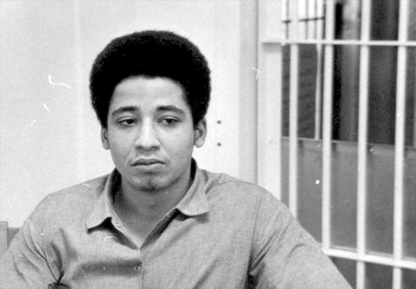A black and white photograph of George Jackson while incarcerated, taken in 1970. He’s seated wearing a collared shirt, looking down and away from the camera in contemplation. Photo Source: African Am