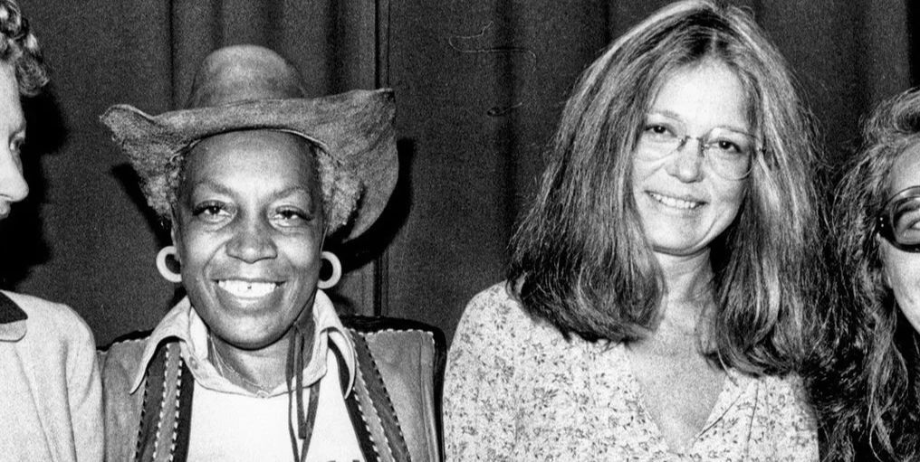 A black and white photograph of Flo Kennedy, left, in cowboy hat and best, T-shirt and hoop earrings and Gloria Steinem, right, in floral patterned top and glasses. Photo Source: New York Public Libra
