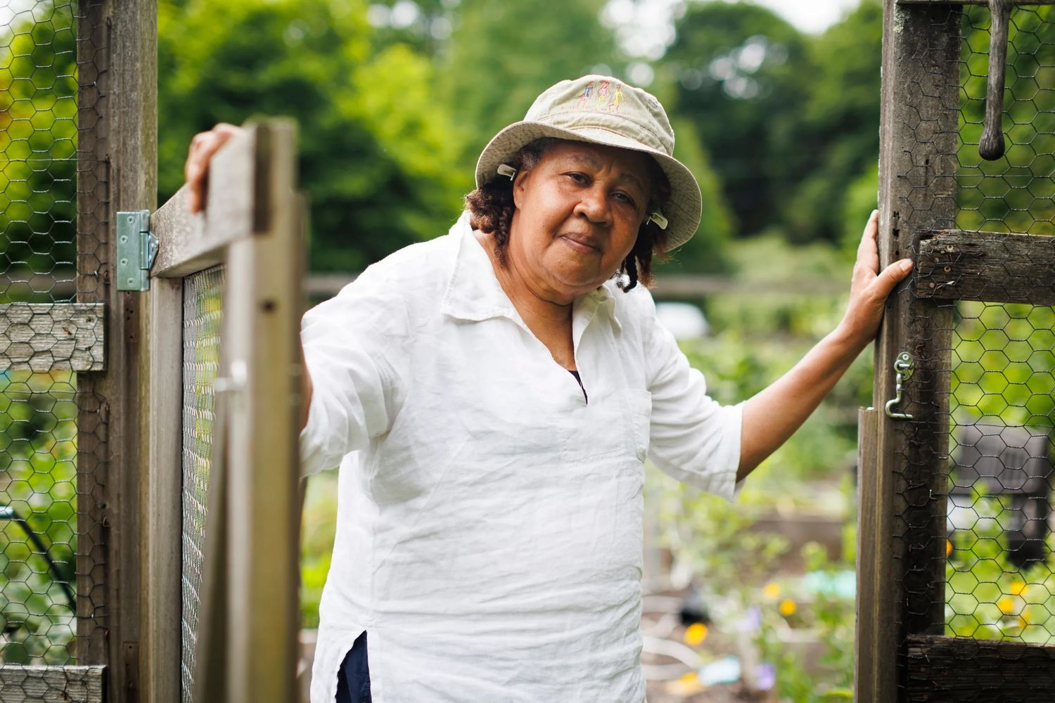 A portrait of Kincaid standing in her gardens, holding a fence door open, wearing a bucket hat and linen blouse, looking at the camera with a friendly gaze. Photo Source: Harvard Gazette.