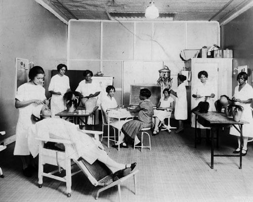 A black and white photograph Joyner (third from right) using her invention on a client while half a dozen other stylists practice on mannequins or people. Photo Source: Library of Congress