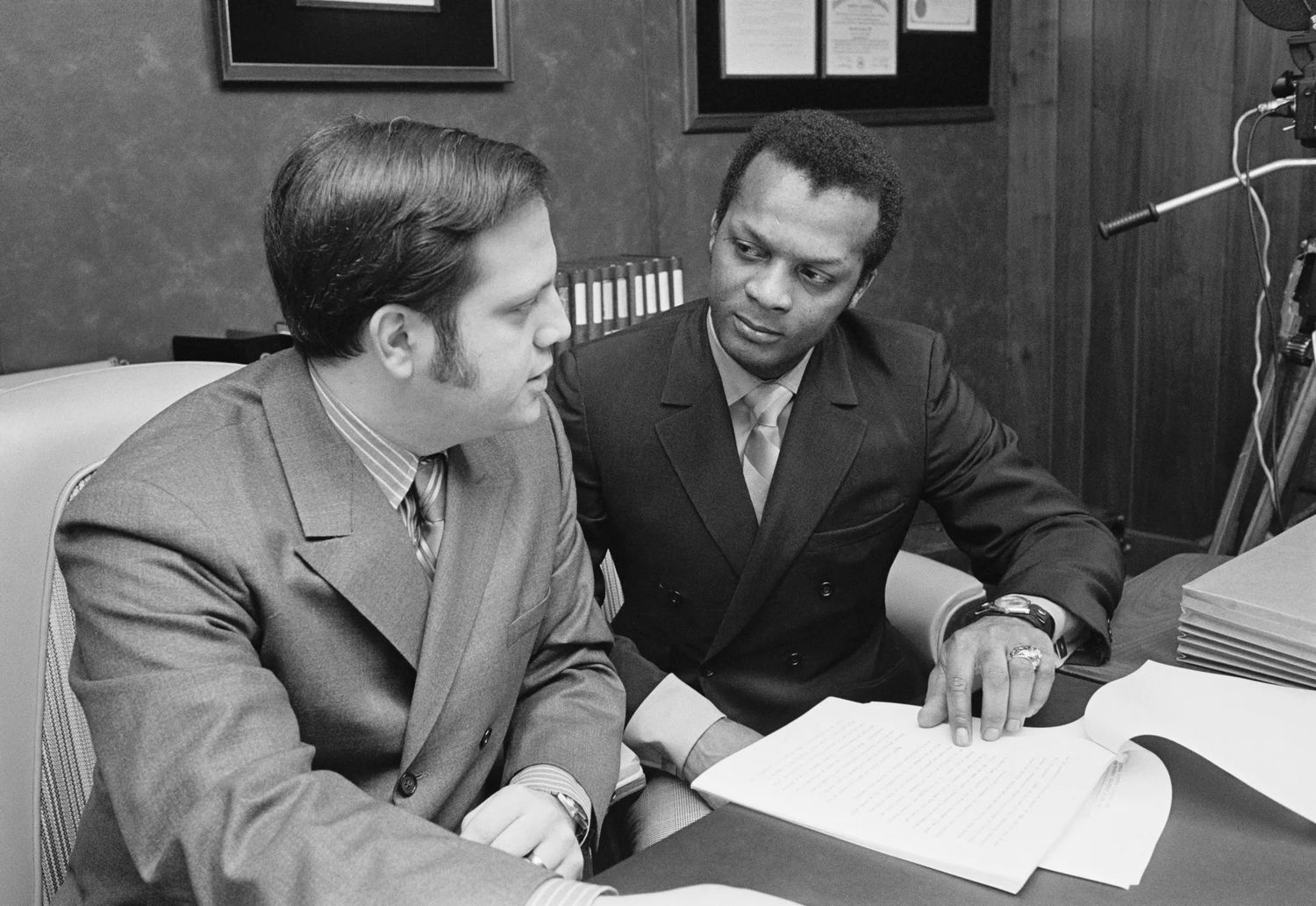 A black and white photo of Curt Flood (right) with his attorney Allan H. Zerman (left) in an office space or court room discussing a stack of papers. Photo Source: Getty Images