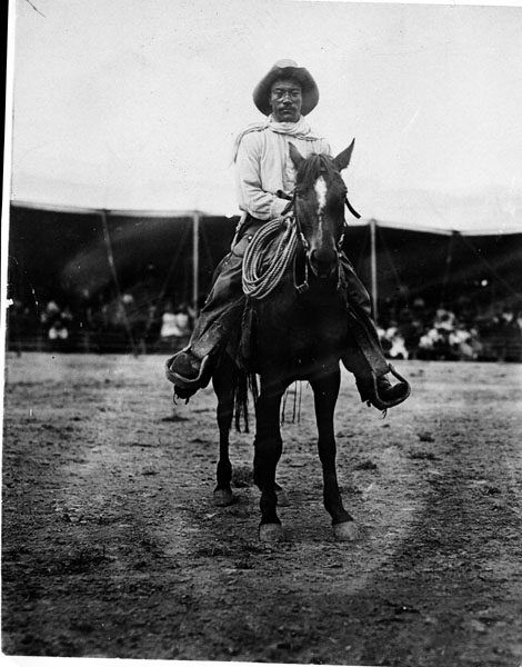 A grainy photograph of Pickett, sitting atop a horse outdoors, in a button down, hat, and chaps, wearing a solemn expression on his face. Photo Source: Tad S. Mizwa Collection, Dickinson Research Cent