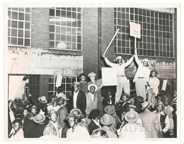 A black and white photograph of the worker strike at R. J. Reynolds Tobacco Company in 1947, depicting dozens of men protesting in front of a building, some hoisted on the shoulders of others. Photo S
