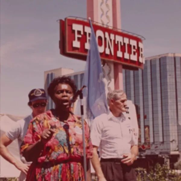 A color photograph of Canty, center, wearing a floral and collared dress, speaking into a microphone on a stage flanked by two men, outside a sign for the Frontier Hotel and Casino. Photo Source: CWA