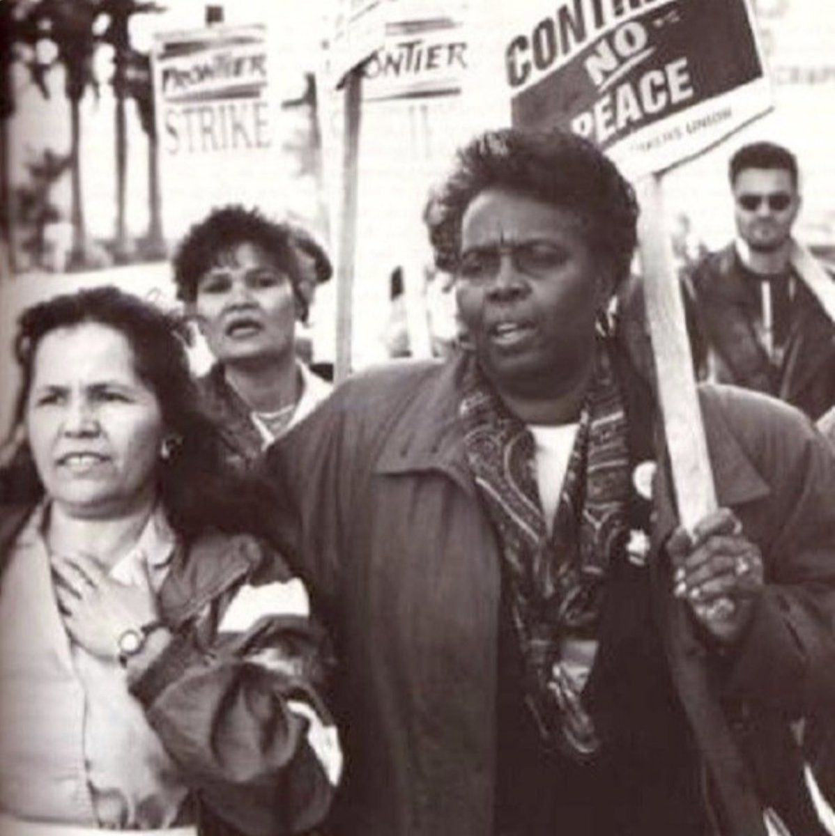 A black and white photograph of Canty, right, holding a strike sign and demonstrating, her arm around another woman, surrounded by other strikers. Photo Source: UAW