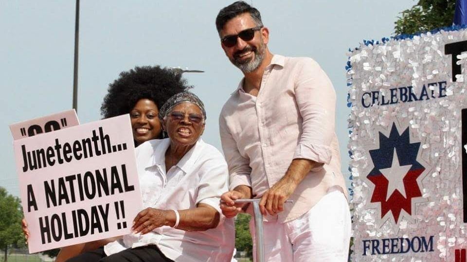 Lee, center, is seated on a float wearing a white shirt and black pants, holding a sign that says “Juneteenth…A NATIONAL HOLIDAY!!” flanked by two other demonstrators. Photo Source: Fort Worth Junetee