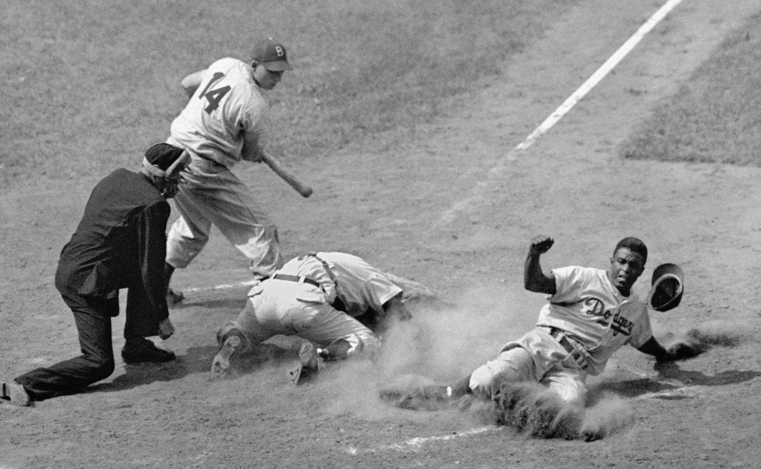 A black and white photograph of Robinson, right, sliding into home base while two players admonish their loss and the umpire watching closely to make a call. Photo Source: Associated Press