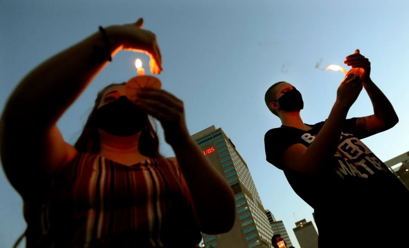 A photograph, taken from below the subjects, depicts two people holding candles and cupping the flame with their hands underneath the evening sky. Photo Source: The Tennessean