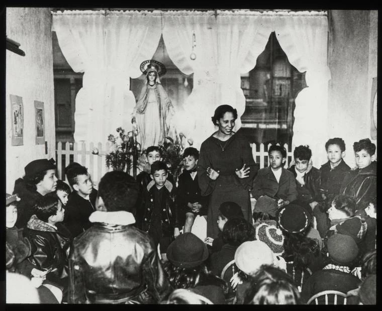 A black and white photograph of Belpré, center, standing with her hands open mid-story, storytelling for dozens of children standing and sitting around her during 115th Street’s storytelling group. Ph